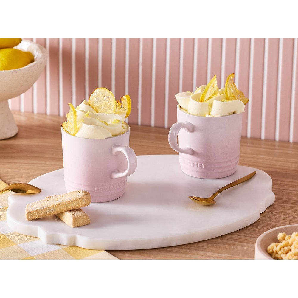 Due tazze rosa con crema e fette di limone su un piatto bianco, accanto a biscotti e un cucchiaino d'oro. Sfondo con strisce rosa.