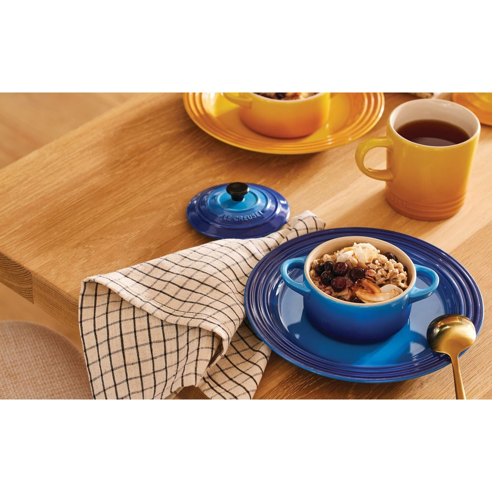Ciotola blu con manici su piatto blu, alimento assortito all'interno, tovagliolo a quadri grigi e bianchi, tazza gialla e tavolo di legno chiaro.