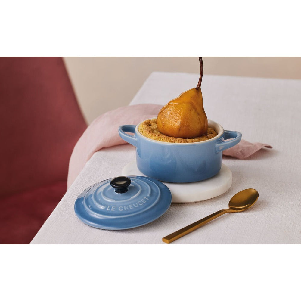Casseruola blu con coperchio, contenente un dolce con una pera, posata d’oro e tovaglia bianca sul tavolo. Sfondo con sedia rosa.