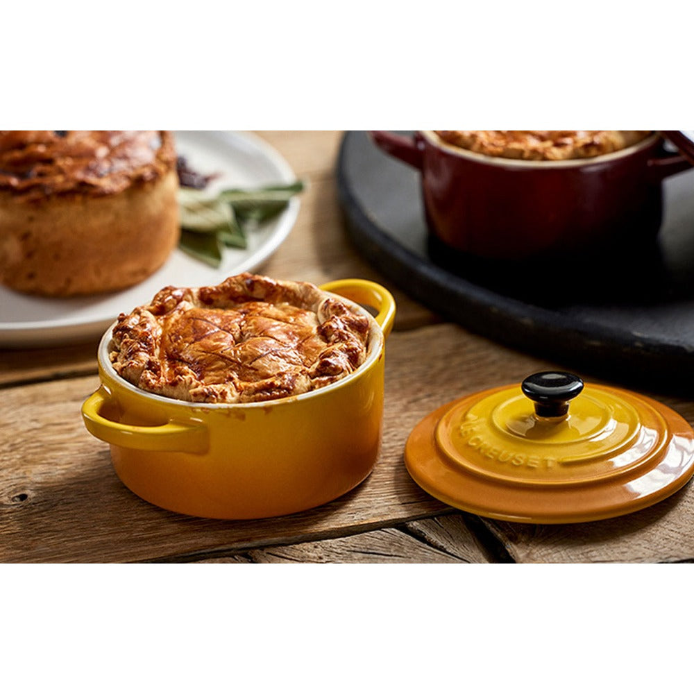 Cocotte gialla con coperchio nero, contenente un piatto dorato. Elementi in legno senza altri oggetti visibili.