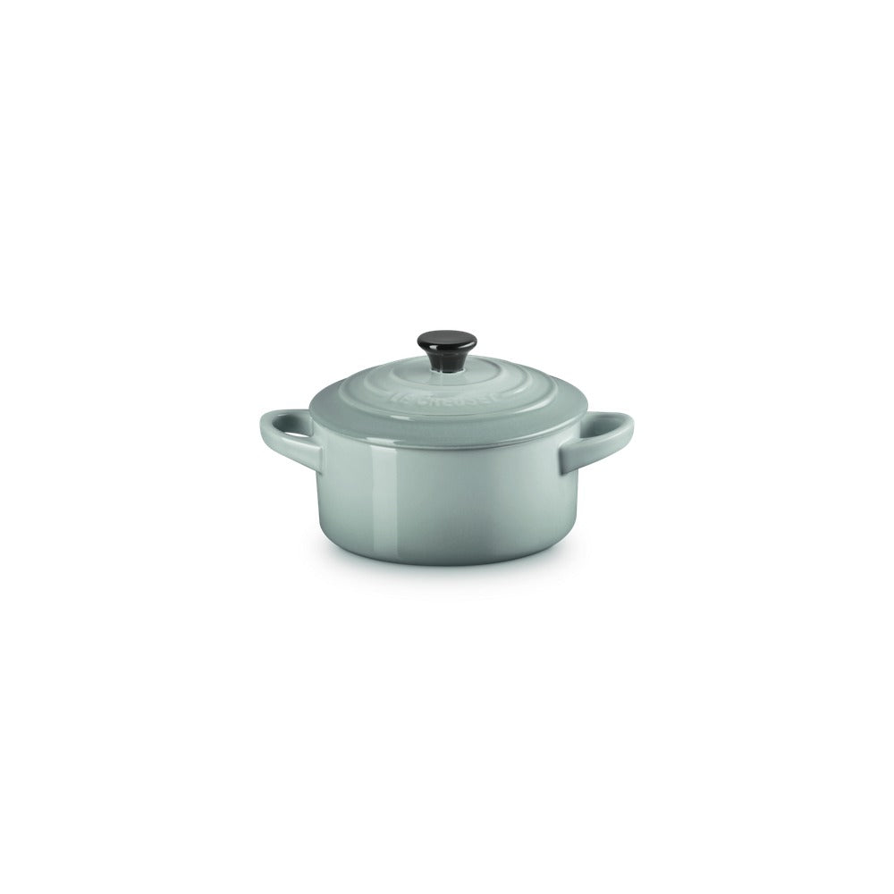 Casseruola rotonda in ceramica verde chiaro con manici, coperchio e pomello nero. Superficie liscia e lucida.