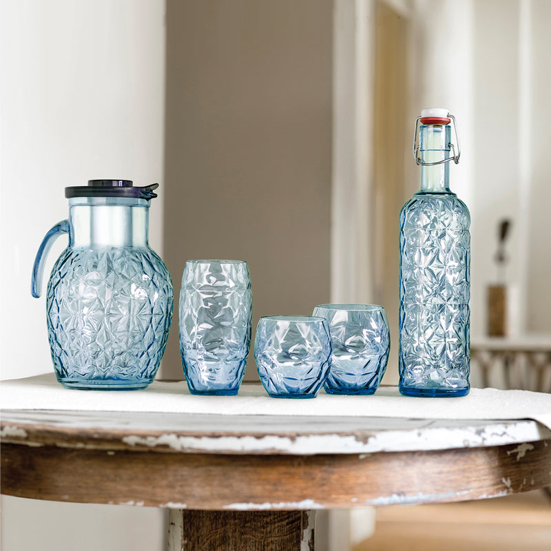 Set de carafe, bouteille et trois verres en verre bleu clair avec motif floral, posés sur table en bois en intérieur.