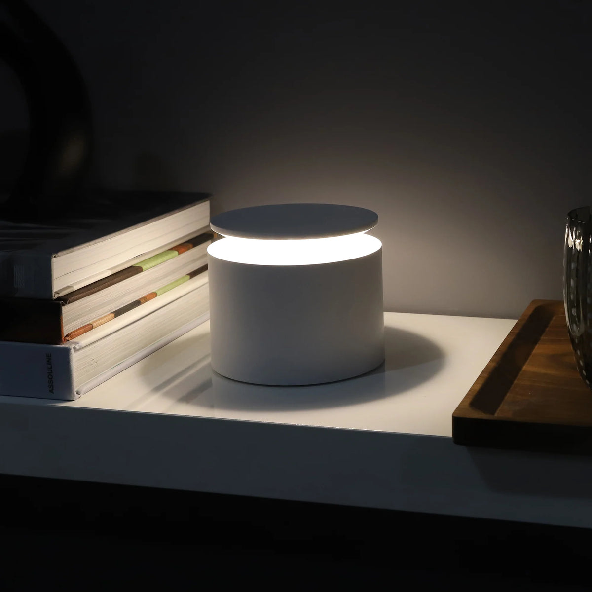 Weiße zylindrische Tischlampe beleuchtet, mit flacher Basis und Abdeckung, auf Regal neben Büchern und Holztablett.