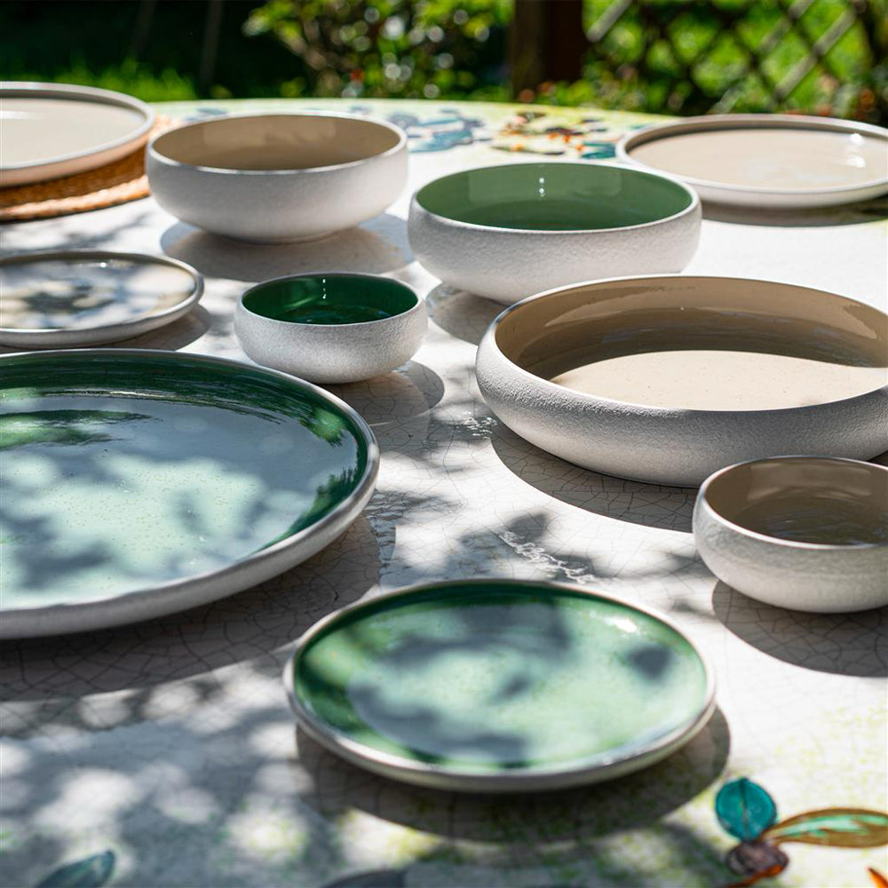 Set di stoviglie in ceramica con piatti e ciotole di varie dimensioni, interni verdi e beige, esterni bianchi, su tavolo all’aperto con ombre di