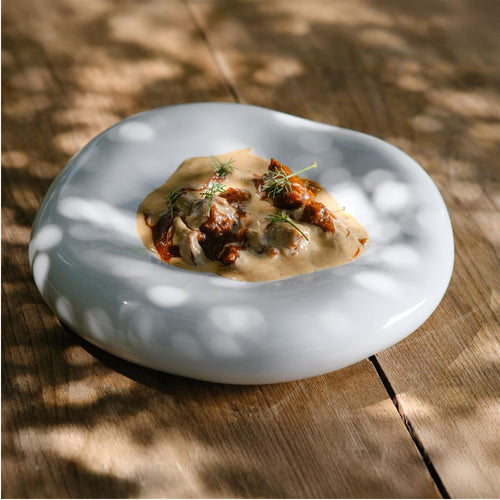 Piatto bianco lucido irregolare con cremoso sugo beige e pezzi di carne rossa guarniti con erbe verdi, su tavolo di legno.