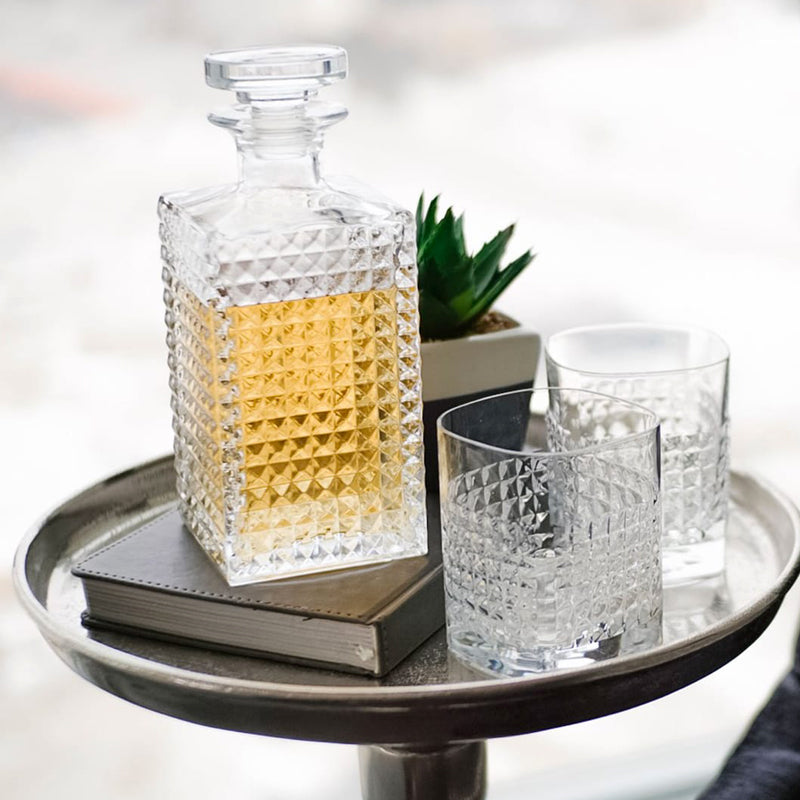 Carafe carrée en verre avec liquide doré, deux verres, livre gris et plante sur caisse métallique ronde