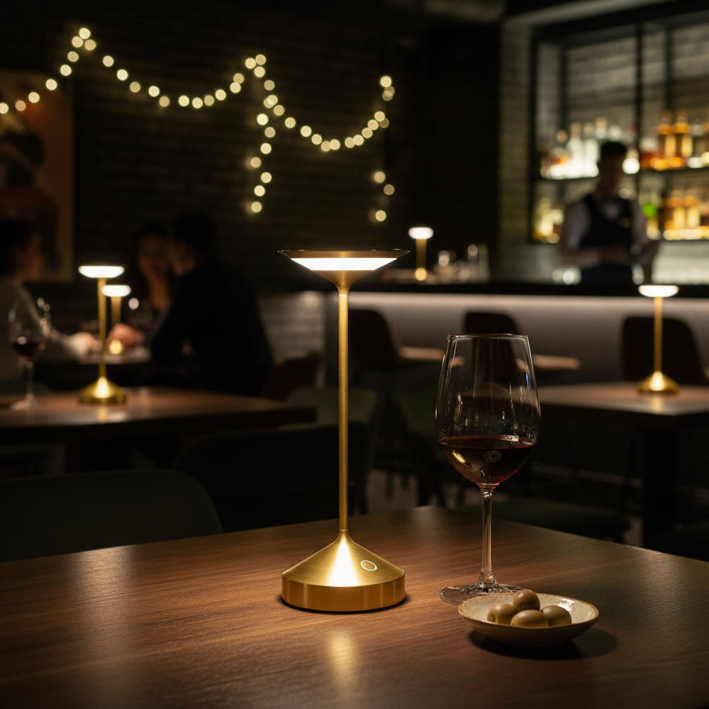 Lámpara de mesa dorada con forma de cáliz iluminado, con copa de vino tinto y cuenco con aceitunas sobre mesa de madera en local con luz tenue.