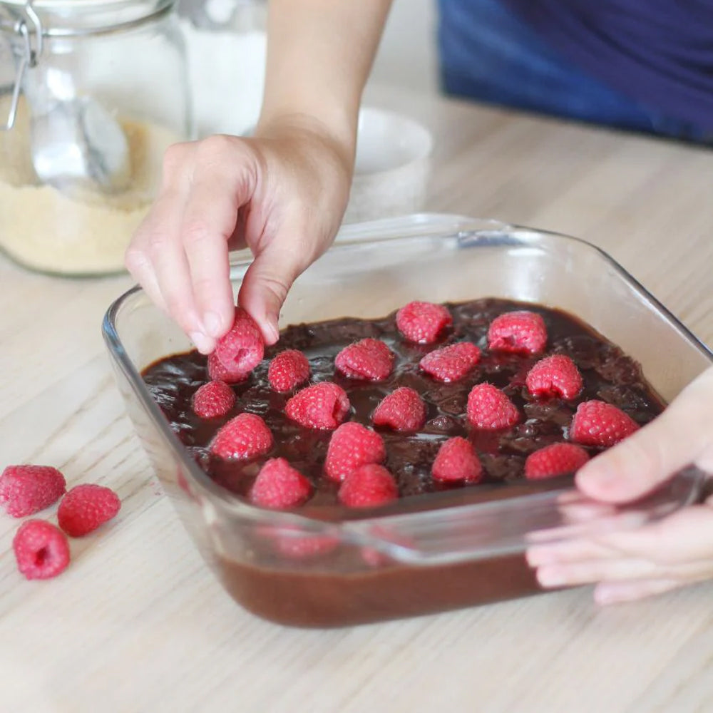 Dessert al cioccolato con lamponi freschi, ciotole e zucchero sullo sfondo, superficie in legno chiaro.