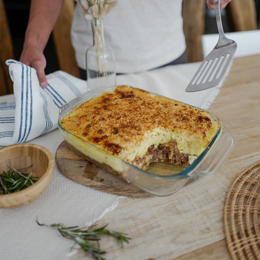 Teglia di lasagna dorata con ripieno, servita su un tagliere di legno, accanto a una ciotola di legno e rametti di rosmarino.