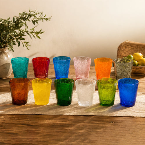 Tredici bicchieri colorati in vetro, disposti su un tavolo di legno, con un vaso di piante a sinistra e un cesto di limoni a destra.