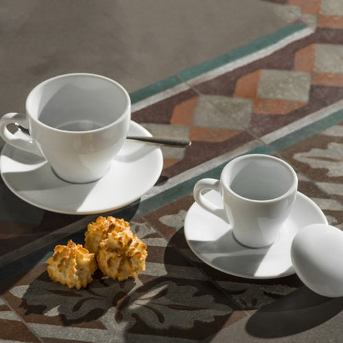 tasse à café et tasse blanches en céramique avec soucoupes, cuillère à café et trois biscuits sur table avec carreaux colorés