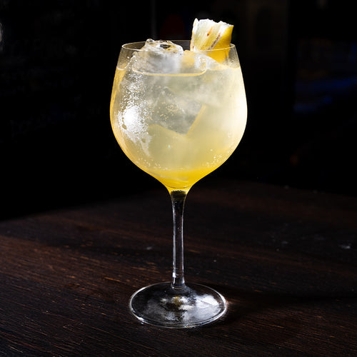 Transparentes Glas mit hellgelbem Cocktail, Eis und Ananasscheibe, auf dunklem Tisch mit schwarzem Hintergrund.