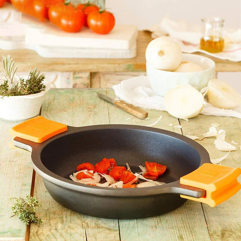 Pentola nera con manici arancioni, contenente pomodori e cipolle. Sullo sfondo, pomodori freschi e cipolle in una ciotola. Ambiente cucina.