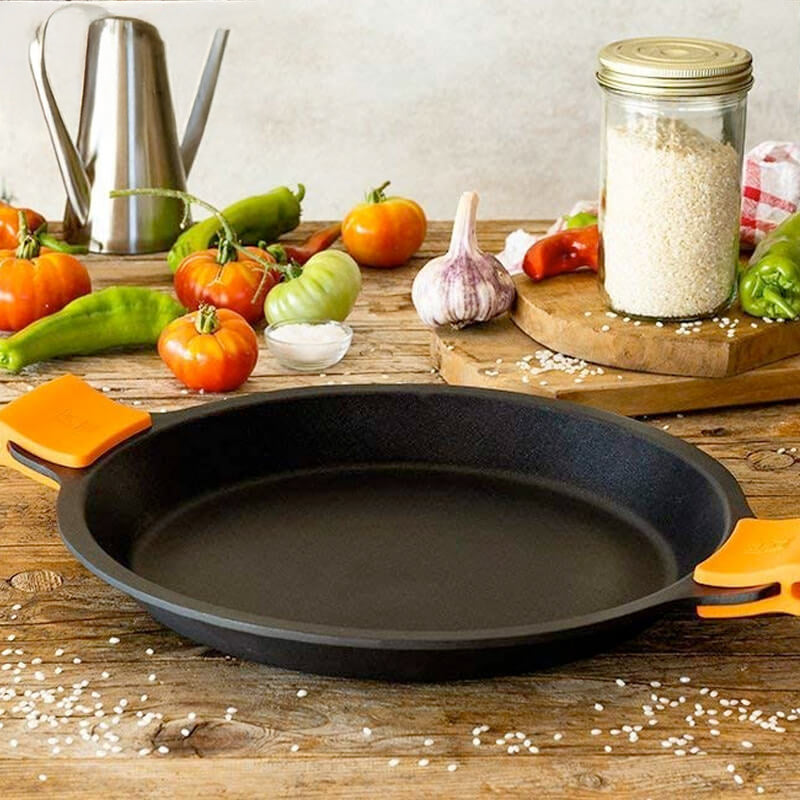 Padella nera con manici arancioni su un tavolo di legno, circondata da pomodori, peperoni, aglio e un barattolo di riso.
