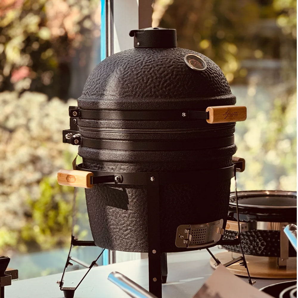 Barbecue noir en céramique avec poignées en bois et thermomètre intégré, posé sur une table près d'une fenêtre avec fond naturel