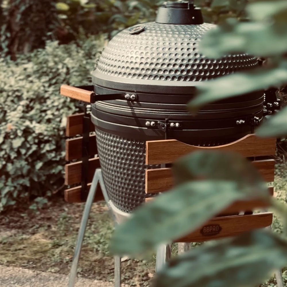 Grauer keramischer eiförmiger Grill mit Holzgriffen, Metallständer und grünen Pflanzen im Hintergrund