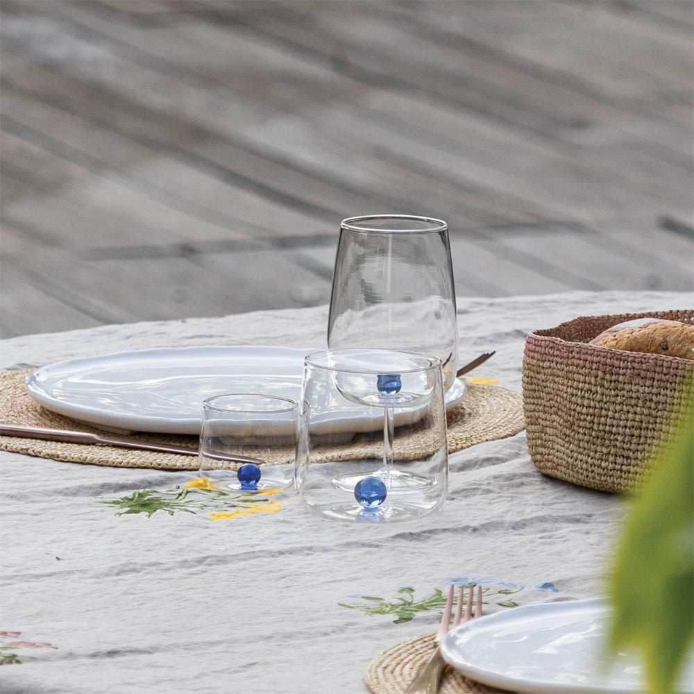 Gedekte tafel met gedecoreerd licht tafelkleed, witte borden op rieten onderborden, drie transparante glazen bekers met blauwe bollen erin