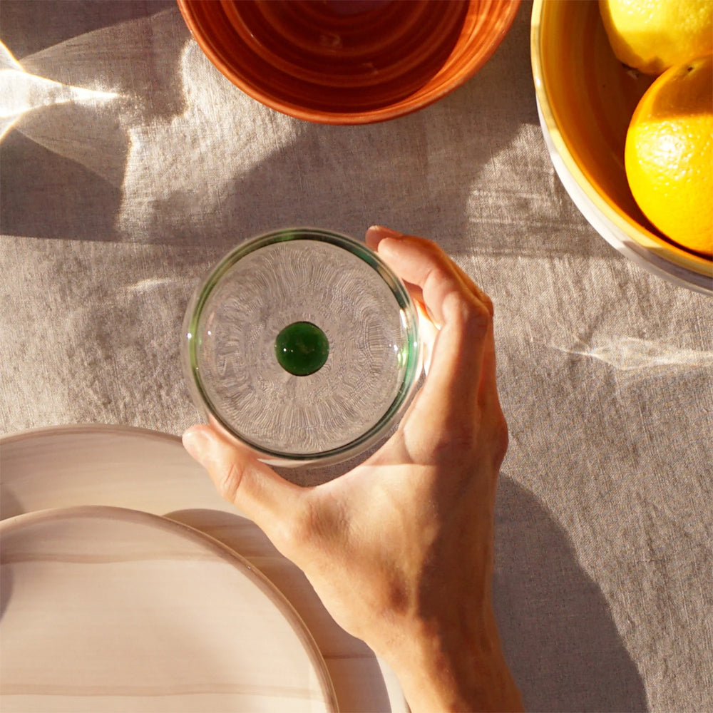Hand houdt een transparant glas met een groene knikker in het midden, beige tafelkleed, borden en oranje kom met sinaasappels.