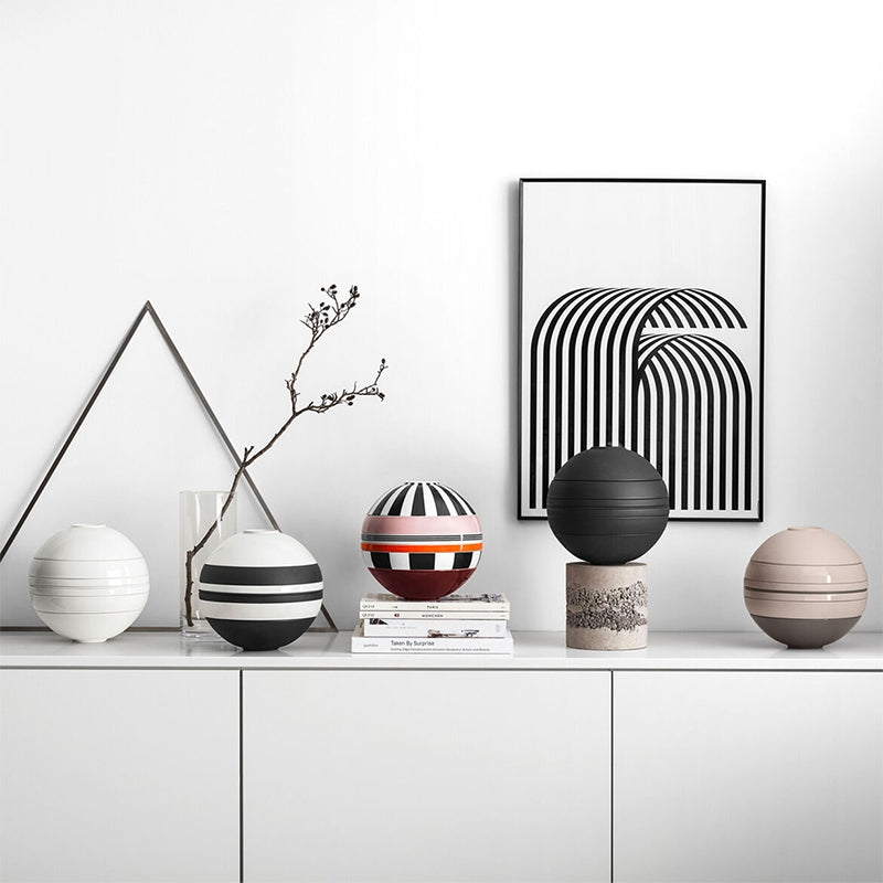 Cinq sphères décoratives en céramique avec rayures horizontales, couleurs blanc, noir, rose et rouge, sur buffet blanc avec tableaux et branche sèche.