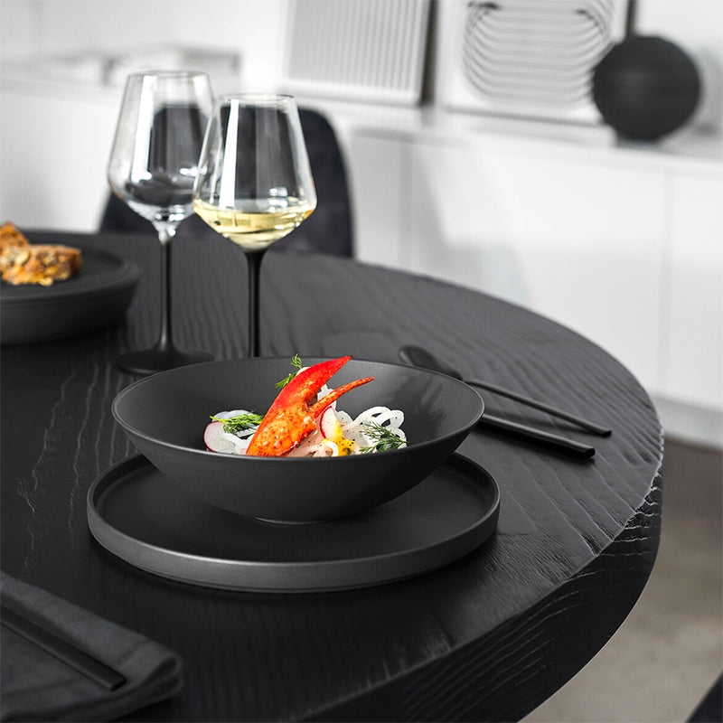 Bol et assiette noirs mats avec salade et chair de crabe rouge sur table ronde noire, deux calices de vin blanc en arrière-plan