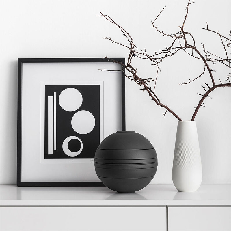Vase sphérique noir mat avec gravures horizontales sur meuble blanc, à côté de vase blanc avec branches sèches et tableau géométrique noir et blanc.