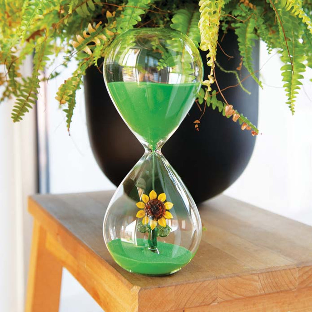 Sablier en verre avec sable vert et tournesol décoratif à l'intérieur, posé sur table en bois devant un vase noir avec fougères vertes.