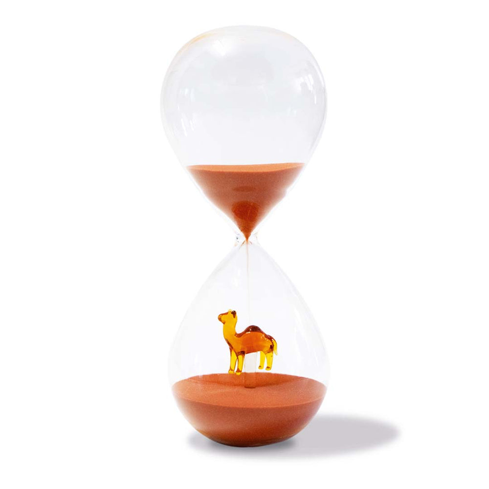 Sablier en verre transparent avec sable rouge et miniature de chameau orange en bas, fond blanc.