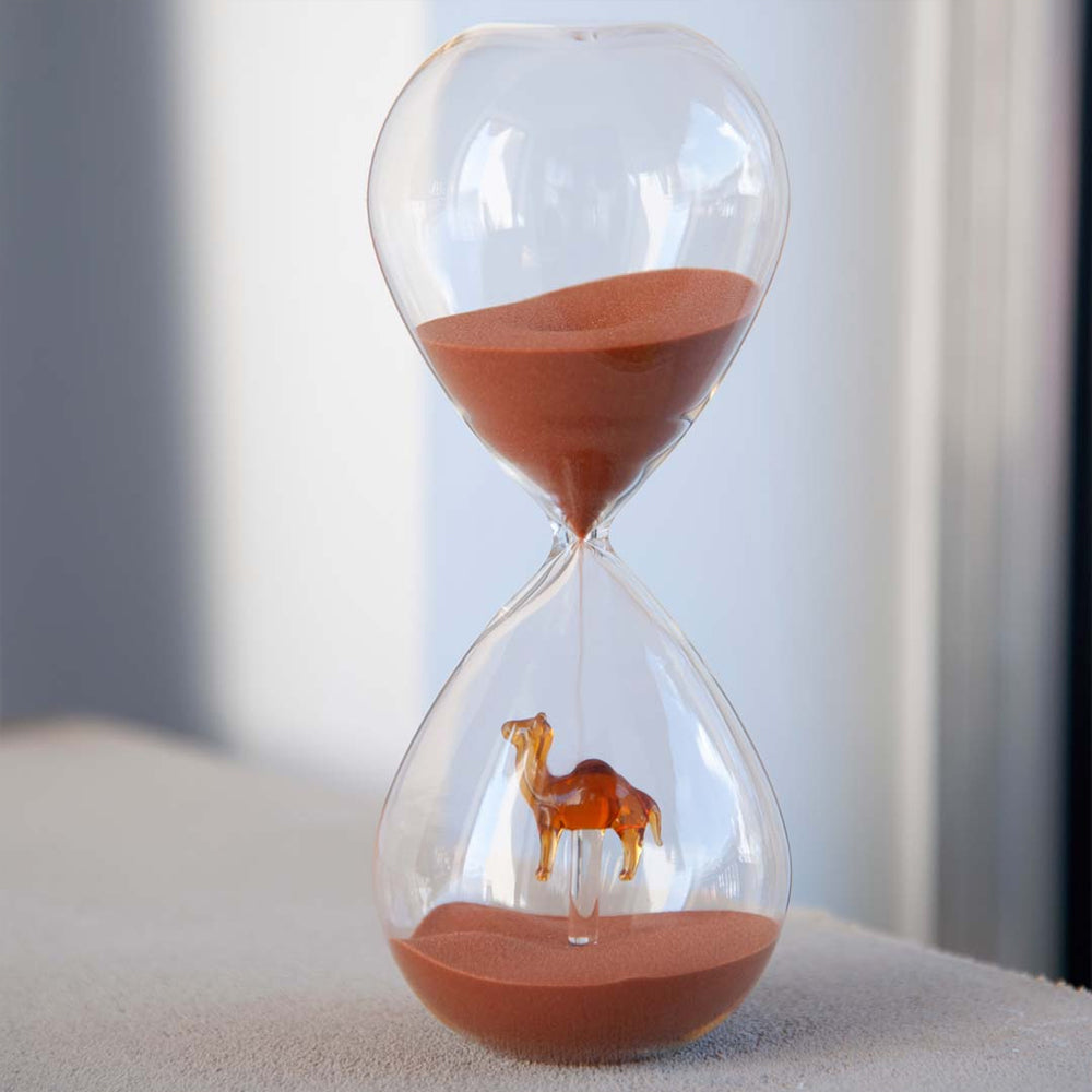 Sablier transparent avec sable rouge et statuette de chameau orange suspendue en bas, sur surface claire.