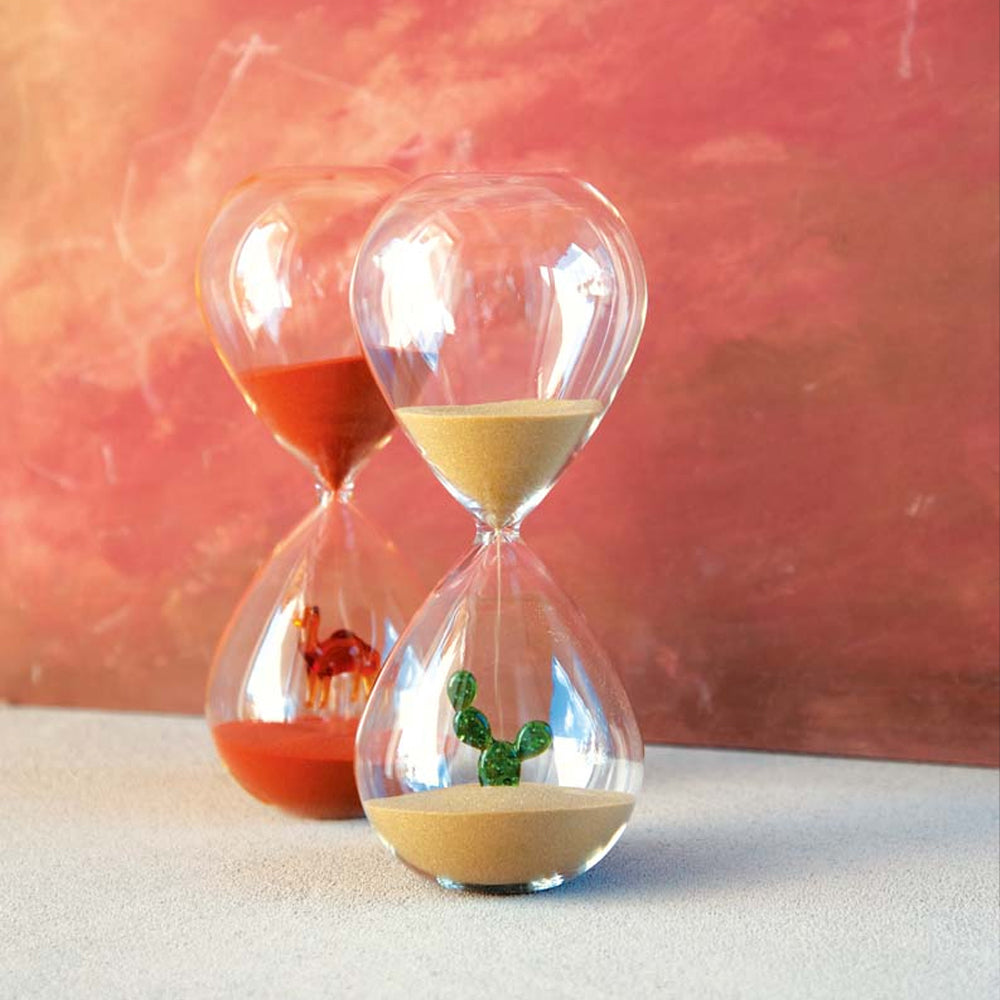 Deux sabliers en verre transparent, un avec sable jaune et cactus vert à l'intérieur, l'autre avec sable rouge et chameau rouge, fond rose.