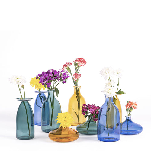 Vasi di vetro colorati in blu, giallo e verde con fiori singoli di vari colori su sfondo bianco.