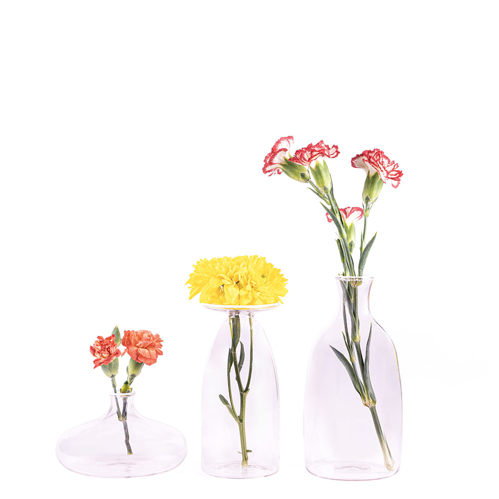 Trois vases en verre transparent avec fleurs : deux petits œillets roses dans vase bas, un chrysanthème jaune dans vase moyen et trois œillets roses dans vase haut