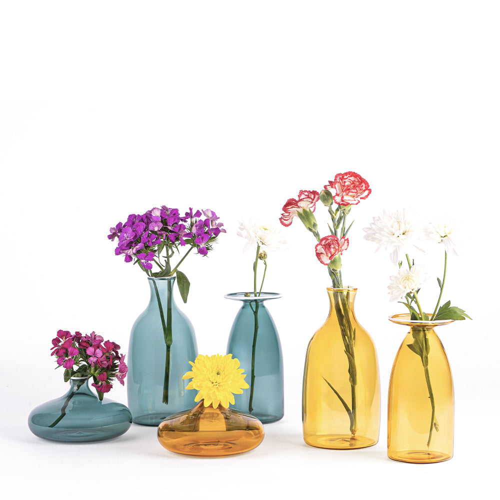 Six vases en verre coloré, bleu et ambré, avec des fleurs de diverses couleurs, sur fond blanc.