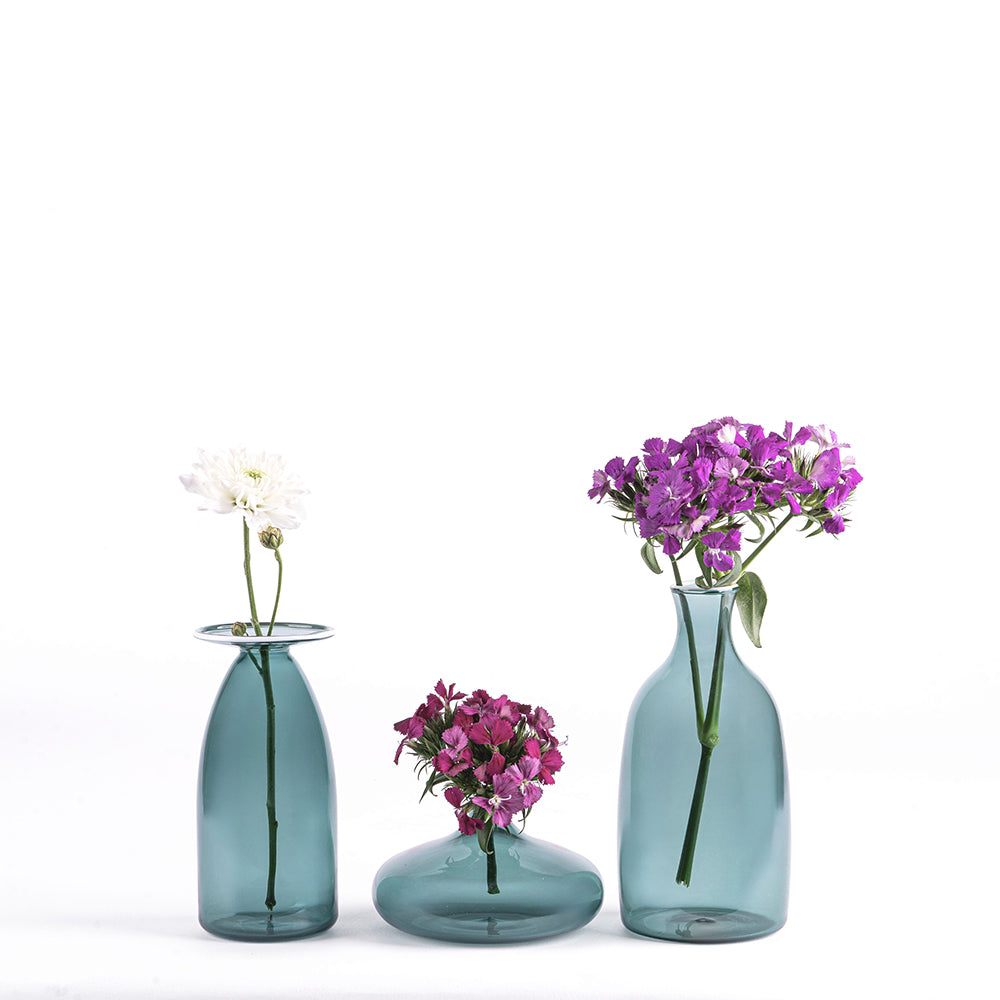 Trois vases en verre vert d'eau avec une fleur blanche, un bouquet de fleurs lilas et un bouquet de fleurs violettes sur fond blanc.