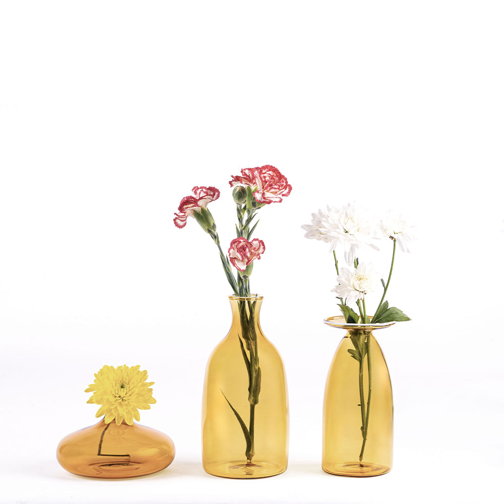 Trois vases en verre ambré de formes différentes, contenant des fleurs : un vase bas avec fleur jaune, un vase moyen avec œillets rouges et un vase haut avec