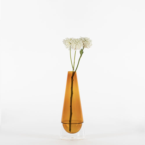 Vaso trasparente con base chiara e corpo ambrato con tre fiori bianchi inseriti su sfondo bianco.