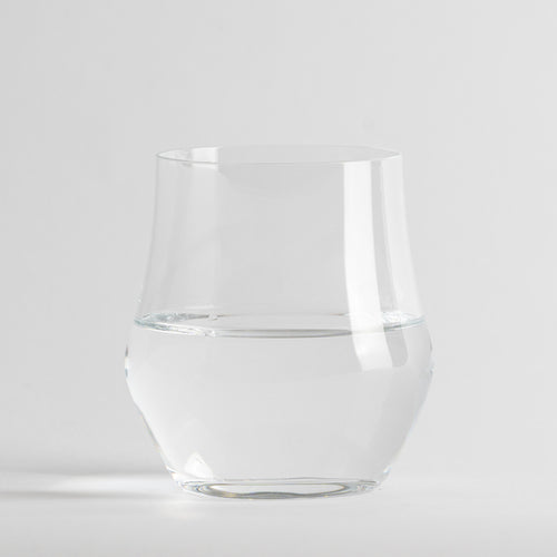 Bicchiere trasparente in vetro contenente acqua, sfondo e superficie bianchi neutri.