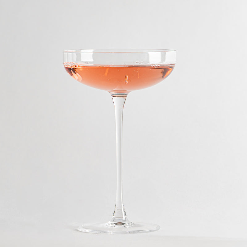 Transparant wijnglas met dunne steel en roze-oranje vloeistof binnenin op witte achtergrond.
