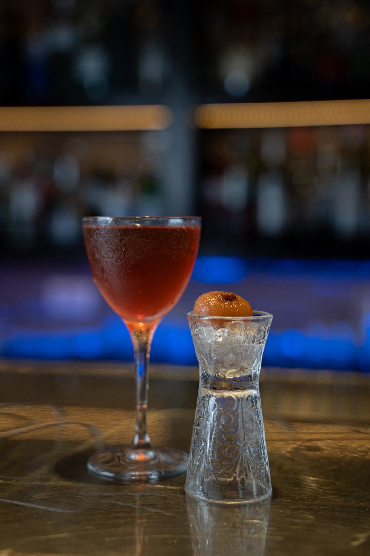 Cocktail rouge dans un verre élégant, à côté d'un verre transparent avec glace et boule orange. Fond flou.