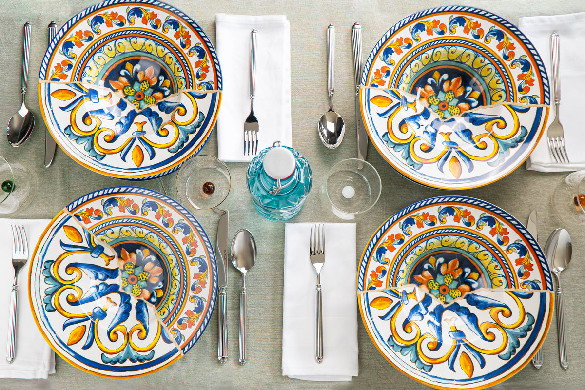 Set of decorated plates with colourful patterns, steel cutlery, transparent glasses and a green water bottle, on light tablecloth.