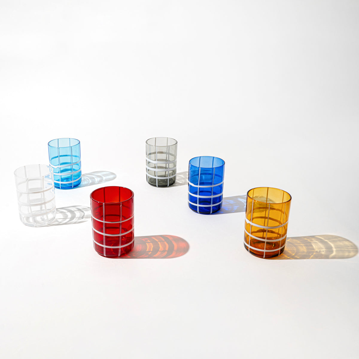 Set di sei bicchieri cilindrici in vetro trasparente colorato con motivo a griglia bianca, su sfondo bianco.