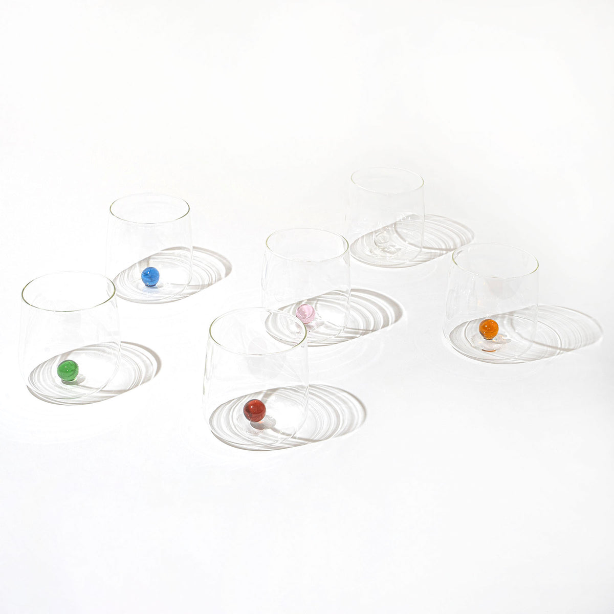 zes transparante glazen bekers ondersteboven met gekleurde knikkers (groen, blauw, roze, oranje, rood) onder elk op witte achtergrond