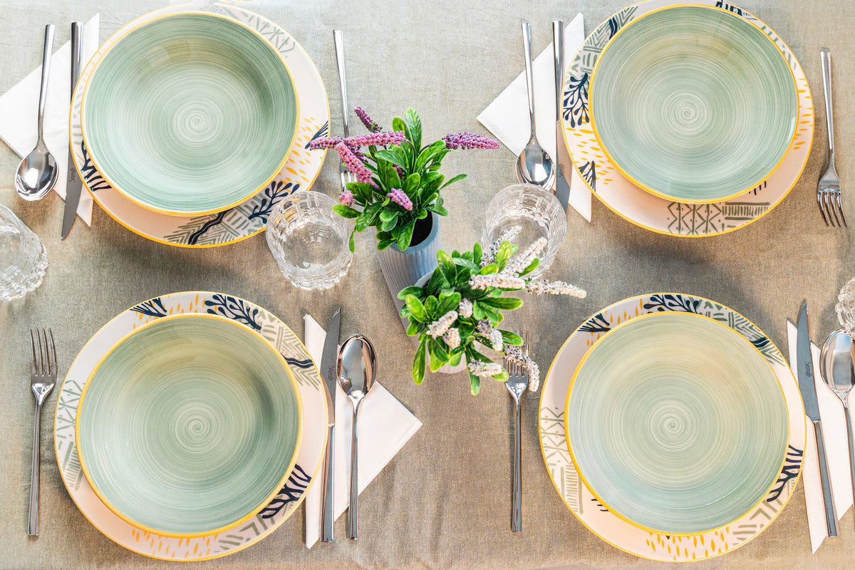 Four decorated ceramic plates, placed on a light tablecloth, with cutlery and transparent glasses. Two small vases with green plants and flowers.