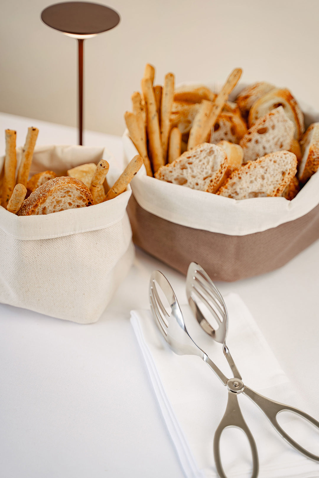 Paniers à pain en tissu avec gressins et tranches de pain ; couverts en argent à côté de serviettes blanches sur fond clair.