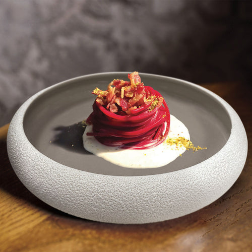 Grauer Teller mit rauem weißem Rand, mit Nest aus roten Spaghetti, weißer Sahne, Pancetta und gelbem Pulver auf Holztisch.