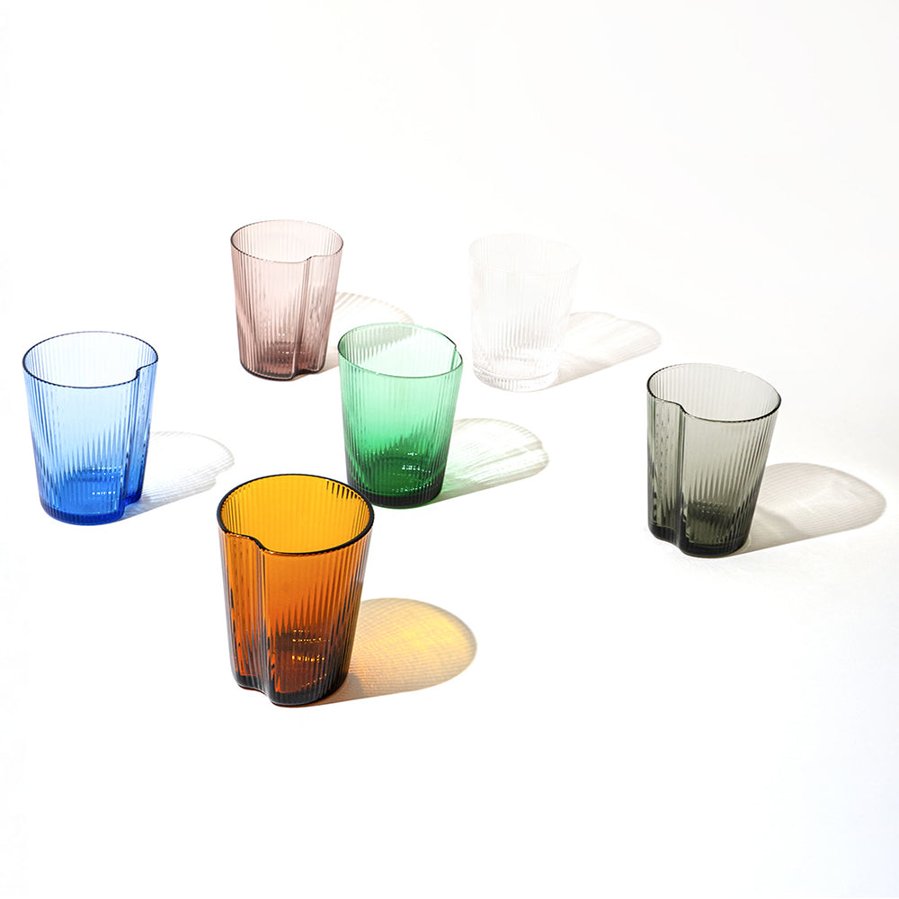 Six verres colorés en verre transparent avec surface cannelée, disposés sur fond blanc avec ombres visibles.