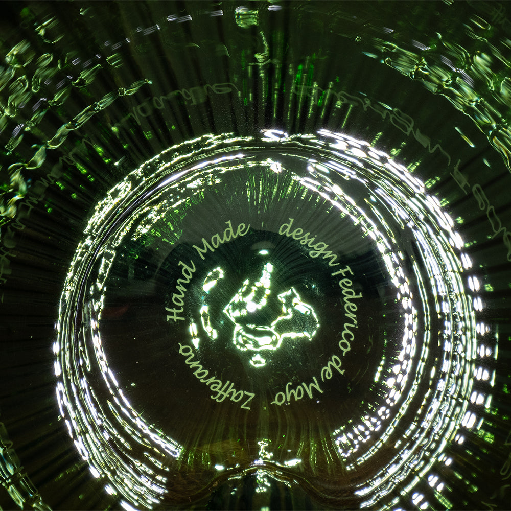 fond d'un objet en verre vert avec inscription circulaire gravée "Hand Made design Federico de Majo Zafferano" et reflets de lumière blanche