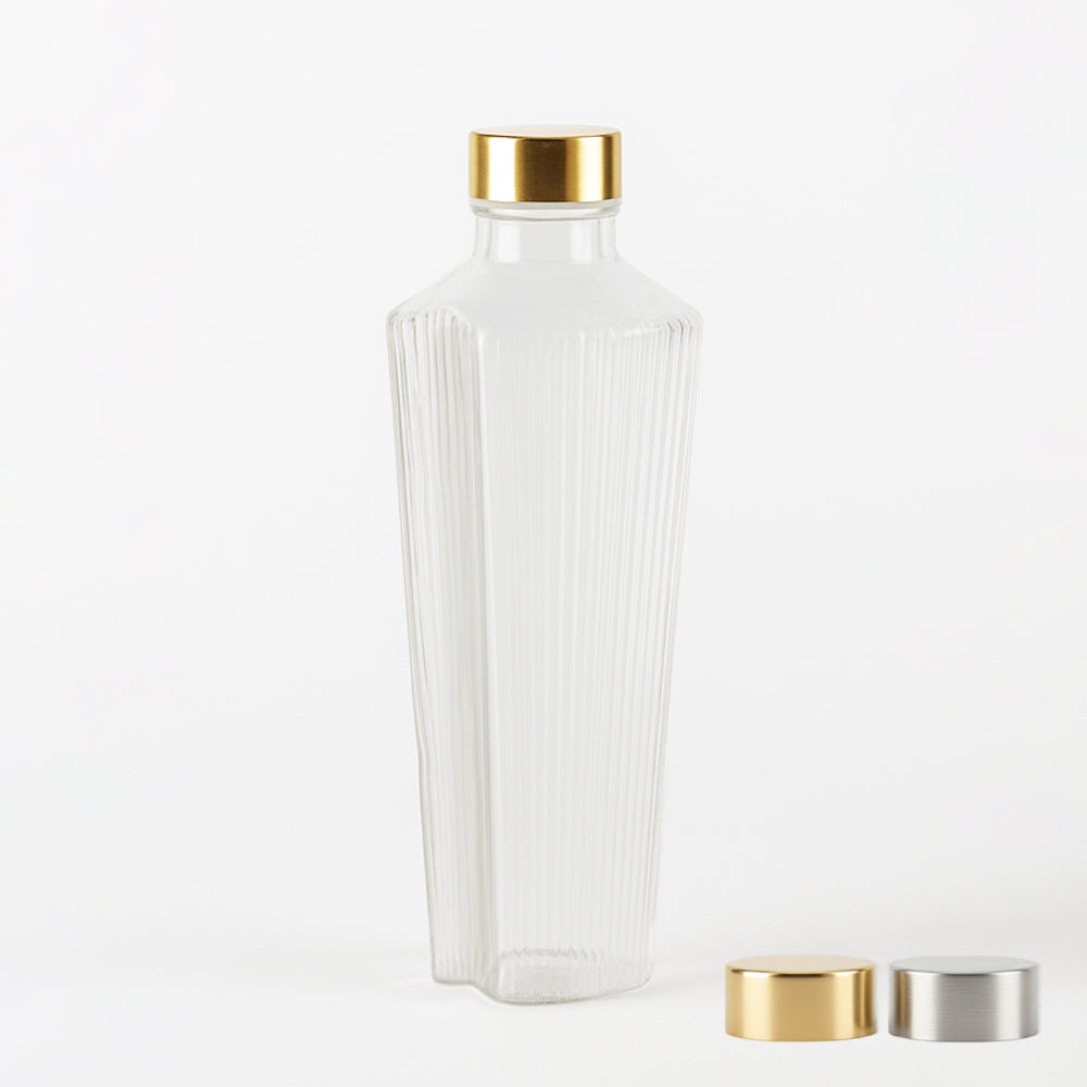 Botella transparente de vidrio con superficies acanaladas verticalmente y tapón dorado, con dos tapones metálicos adicionales, dorado y plateado, al fondo