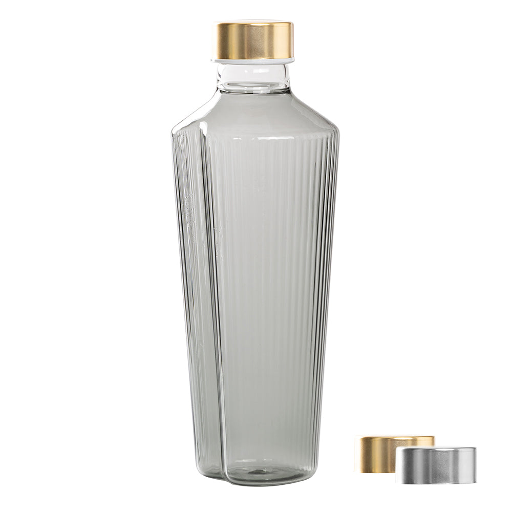 Botella de vidrio transparente con base hexagonal y estrías verticales, tapón dorado y dos tapones metálicos, uno dorado y uno plateado.