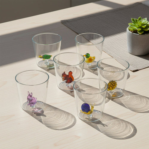 Sei bicchieri trasparenti in vetro con piccole sculture di animali colorati sul fondo, su tavolo chiaro, pianta a destra.