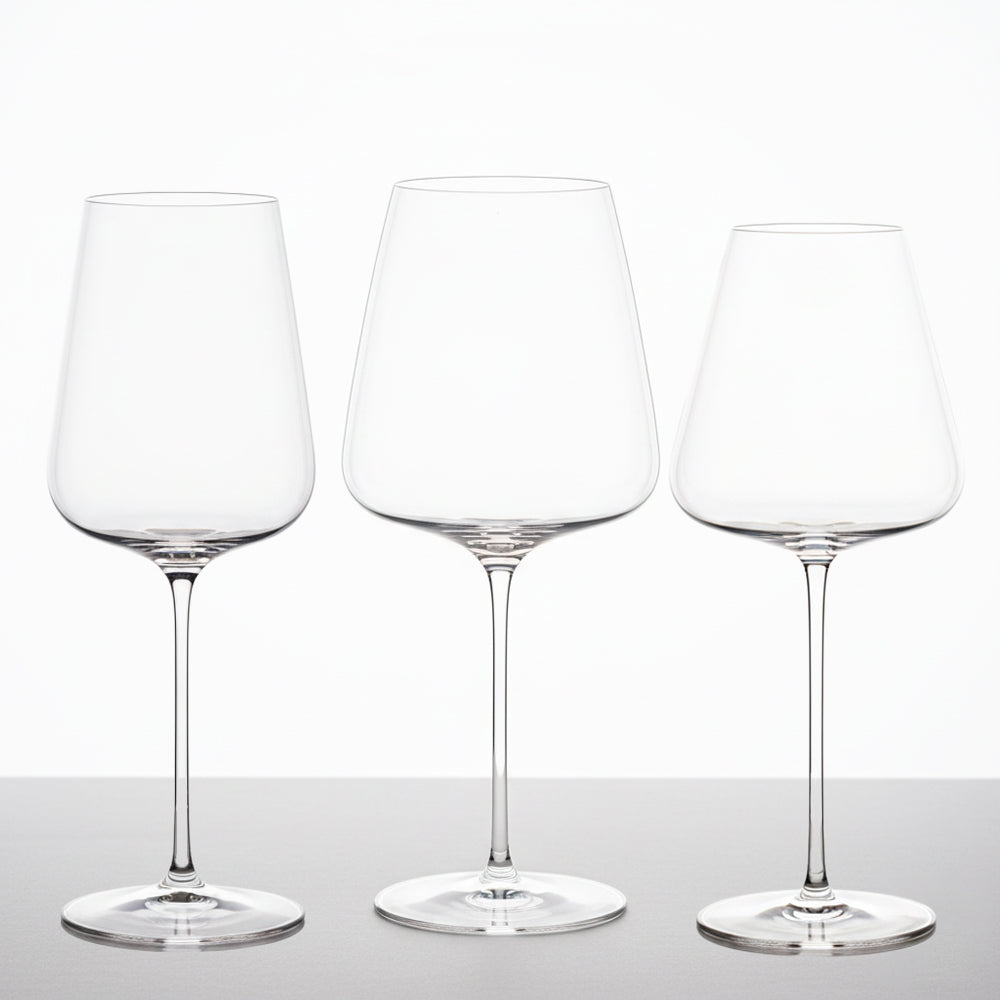 Three transparent glass goblets with thin stem arranged on grey surface and neutral white background.